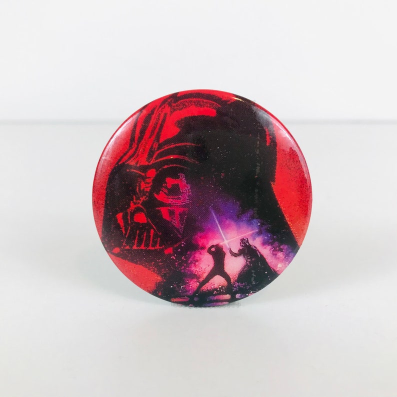 May include: A red, white, and black button featuring a silhouette of Darth Vader from Star Wars. The button shows a scene of Darth Vader and Luke Skywalker fighting with lightsabers.