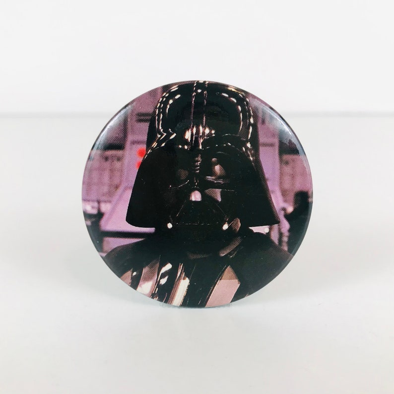May include: A round button featuring a black and white image of Darth Vader from Star Wars. The button is a collectible for fans of the Star Wars franchise.