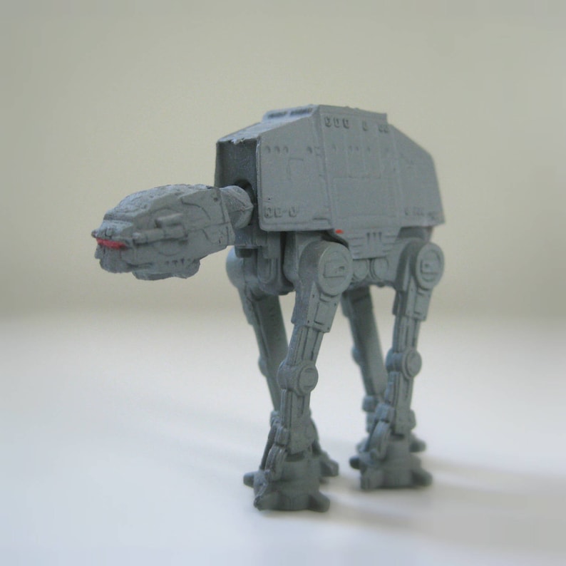 May include: A gray plastic toy model of an AT-AT walker from the Star Wars franchise. The model is detailed with articulated legs and a raised platform.