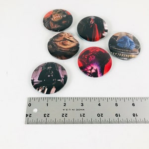 May include: Six round Star Wars themed buttons featuring characters from the movie franchise. The buttons feature Darth Vader, Jabba the Hutt, a blue creature, and a red-robed figure. The buttons are approximately 1 inch in diameter.