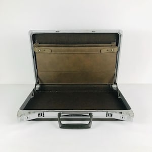 May include: An open, silver-framed briefcase with a black interior and a brown leather pocket. The briefcase has a black handle and is set against a white background. The letters "J" and "K" are visible on the sides.