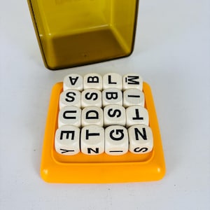 May include: A vintage word game with sixteen white letter dice arranged in a yellow tray. The dice have black letters. A yellow metal box is in the background. The game is designed for word creation and vocabulary building.