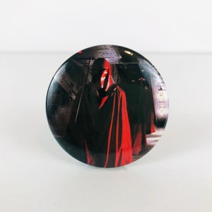 May include: A round button with a picture of a person wearing a red hooded robe and a helmet. The person is standing in a dark, futuristic setting.