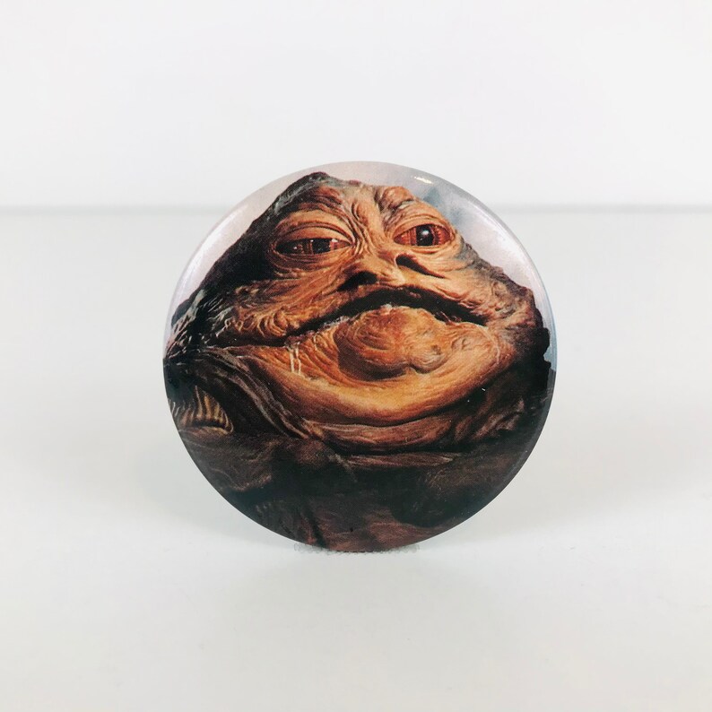 May include: A round button with a picture of Jabba the Hutt from Star Wars. Jabba is a large, brown, slug-like alien with a wide mouth and large eyes.