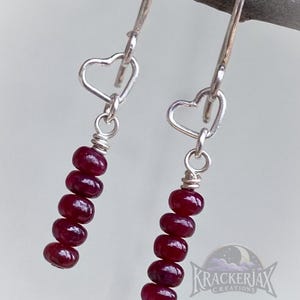 May include: Silver dangle earrings with heart-shaped accents. Each earring features a series of six deep red, oval-shaped beads. The earrings are suspended from a silver hook.