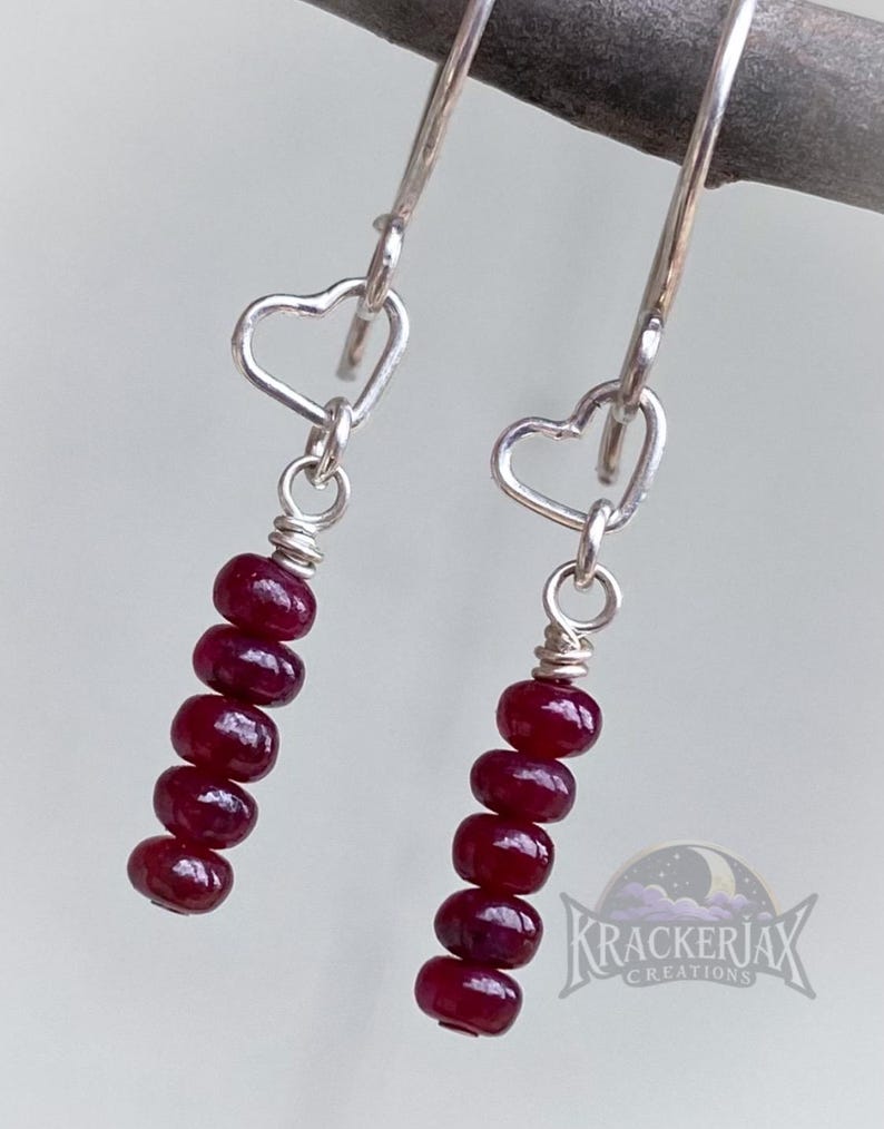 May include: Silver dangle earrings with heart-shaped accents. Each earring features a series of six deep red, oval-shaped beads. The earrings are suspended from a silver hook.