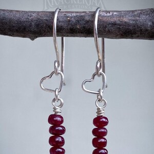 May include: A pair of dangle earrings with silver-toned hooks and heart-shaped accents. Each earring features a strand of six deep red, round beads. The earrings are suspended from a dark brown branch against a light gray background. The brand name "KRACKERJAX CREATIONS" is visible.
