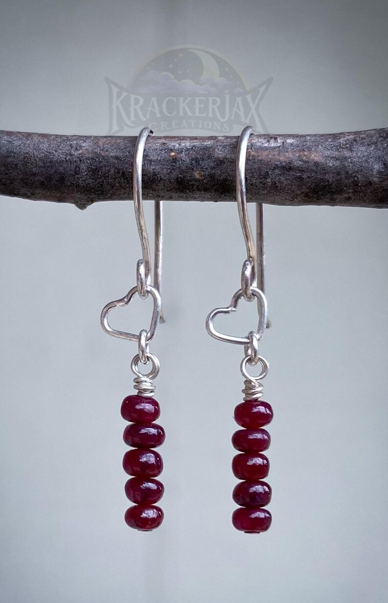 May include: A pair of dangle earrings with silver-toned hooks and heart-shaped accents. Each earring features a strand of six deep red, round beads. The earrings are suspended from a dark brown branch against a light gray background. The brand name "KRACKERJAX CREATIONS" is visible.