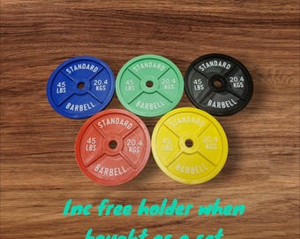 Puede incluir: Cinco placas de peso de barra estándar en diferentes colores: azul, verde, negro, rojo y amarillo. Cada placa pesa 45 libras (20,4 kg). El texto "Inc free holder when bought as a set" está escrito debajo de las placas.