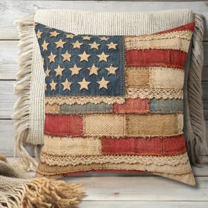 May include: Square decorative pillow with a patchwork design resembling the American flag. The pillow features a blue denim section with star cutouts, red and beige fabric patches, and lace trim. The pillow is placed on a neutral-colored background.