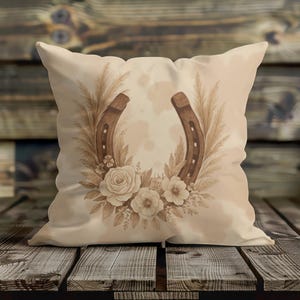 May include: A decorative pillow featuring a horseshoe design. The horseshoe is surrounded by floral and wheat elements in shades of brown and beige. The pillow is placed on a wooden surface.