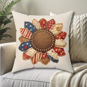 May include: A decorative throw pillow with a sunflower design. The sunflower petals are made of red, white, and blue fabric patches, some with stars and stripes. The center is a brown burlap circle, and the pillow has a cream-colored background.