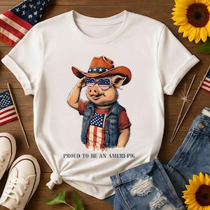 May include: White t-shirt with a graphic of a pig wearing a cowboy hat, sunglasses, and a denim vest with an American flag design. The text "PROUD TO BE AN AMERI-PIG" is below the graphic. The image includes sunflowers, flags, and denim.