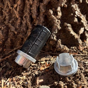 Puede incluir: Un contenedor de metal negro y plateado con una tapa texturizada. El contenedor está abierto y la tapa está tirada en el suelo junto a él.