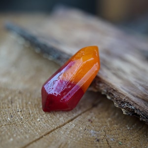 May include: A faceted, gemstone-like object with a gradient of colors, transitioning from deep red at the base to vibrant orange at the top. The object is displayed on a textured wooden surface, showcasing its polished, reflective surface.