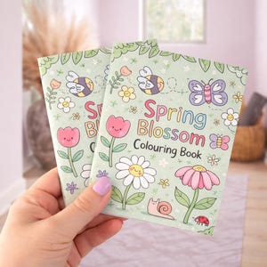 Puede incluir: Dos libros para colorear Spring Blossom con una cubierta verde claro. La cubierta presenta ilustraciones de flores, abejas, mariposas y el texto "Spring Blossom Colouring Book". Los libros son sostenidos por una persona.