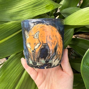 May include: A ceramic mug with a hand-painted orange fox design against a dark blue background. The fox is depicted in a detailed, artistic style. The mug is held in front of lush green foliage.