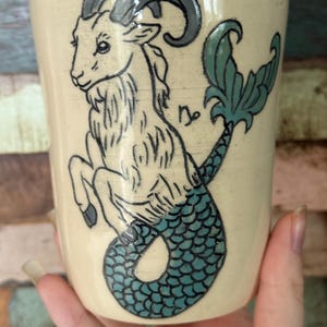 May include: A cream-colored ceramic mug featuring a Capricorn design. The illustration depicts a goat with a fish tail, in shades of black and teal. The mug is held by a person.