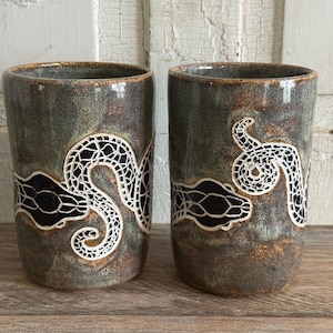 May include: Two cylindrical ceramic tumblers featuring a mottled green and brown glaze. Each cup displays a prominent black snake motif with a white, intricate, lace-like pattern detailing the body and head, wrapping around the exterior surface.