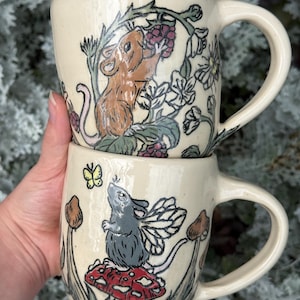 May include: Two cream-colored ceramic mugs stacked vertically. The top mug features a brown mouse eating blackberries among green leaves. The bottom mug displays a gray mouse with wings sitting on a red mushroom with white spots, accompanied by a yellow butterfly.
