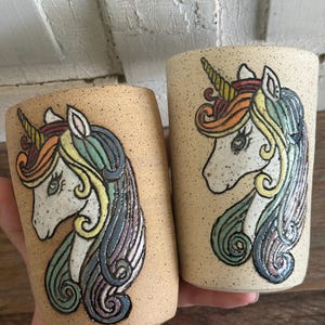 May include: Two cylindrical ceramic cups with a speckled beige finish, each featuring a raised, hand-painted unicorn design. The unicorns have multicolored manes in shades of orange, yellow, green, and purple, with a prominent spiral horn.
