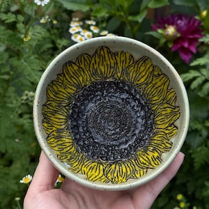 May include: A small, handmade ceramic bowl featuring a sunflower design. The bowl's interior showcases a detailed sunflower with yellow petals and a dark center. The bowl is held in a hand, with a natural background of flowers and greenery.