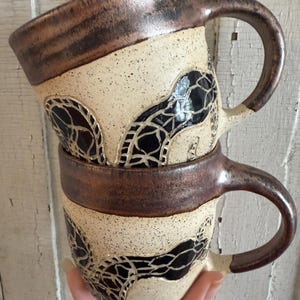 May include: Two handmade ceramic mugs stacked. The mugs have a speckled cream-colored base with a dark brown rim and handle. A black, web-like design decorates the body of each mug. The mugs are held in a hand.