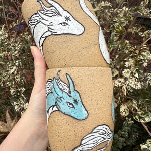 May include: Two beige ceramic tumblers with hand-painted dragon designs. One dragon is light blue, the other is white. The dragons have detailed features and are set against a speckled background. The tumblers are cylindrical and appear to be handmade.