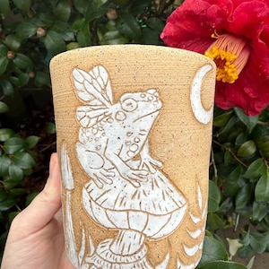 May include: Tan ceramic mug with a white relief design of a frog with fairy wings sitting on a mushroom. A crescent moon is also part of the design. The mug is cylindrical and has a textured surface.