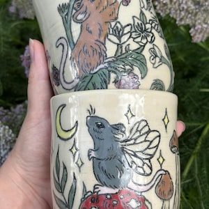May include: Two ceramic mugs with whimsical illustrations. One mug features a brown mouse reaching for berries, the other a gray mouse with fairy wings on a red mushroom. Both mugs have a cream-colored background with floral and celestial details.