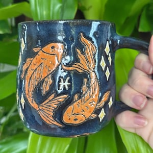 May include: A handmade ceramic mug with a dark blue glaze. The mug features two orange koi fish and a Pisces symbol, with star-shaped accents. The handle is dark blue. The mug is held against a background of green leaves.