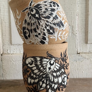 May include: Two stacked ceramic tumblers made of speckled tan clay. Each tumbler features a hand-carved, black and white relief design of a moth surrounded by leafy branches. The moth wings are detailed with crescent moon and star motifs.