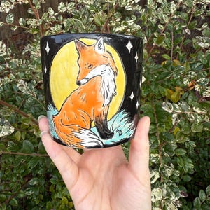 May include: A black ceramic mug featuring a hand-painted fox design. The fox is orange and white, set against a yellow moon and a starry black background. The mug is held in a hand, with green foliage in the background.