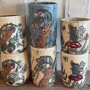 May include: Five ceramic tumblers featuring hand-painted illustrations of mice. Two cups show brown mice with blackberries and bees, two show brown mice with lily of the valley flowers, and two show grey mice with wings perched on red mushrooms.