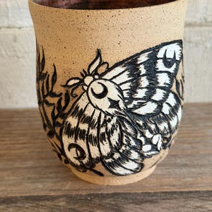May include: A speckled tan ceramic mug featuring a carved and painted black and white moth design on the exterior. The moth has detailed wing patterns and a crescent moon symbol on its body. The interior rim of the mug has a dark brown glaze.