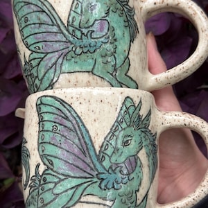 May include: Two speckled ceramic mugs with a teal and purple dragon design. The dragon has large, detailed wings and is set against a cream-colored background. The mugs have large handles.