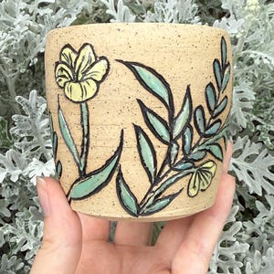 May include: A small, beige ceramic planter with a hand-painted floral design. The planter features a yellow flower and green leaves outlined in black. The pot is held in a hand, with a backdrop of silvery-green foliage.