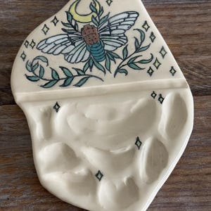 May include: Cream-colored ceramic paint palette with a whimsical design. The top section features a detailed illustration of a winged insect, a crescent moon, and stars. The lower section has several wells for holding paint. Decorative and functional.