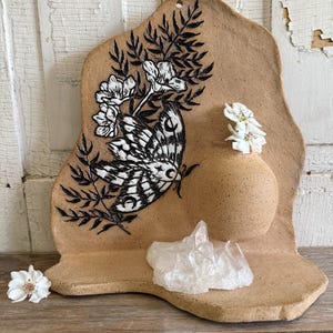 May include: A tan ceramic wall hanging with a moth and floral design. The moth is black and white, and the flowers are white. A small vase holds white flowers, and a clear crystal sits on the shelf. The piece is on a wooden surface.