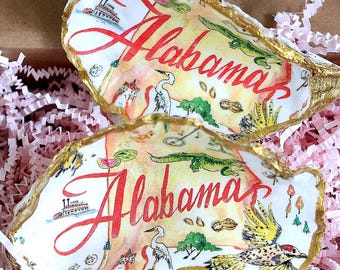 Può includere: Un set di quattro conchiglie di ostrica, ciascuna dipinta a mano con la parola "Alabama" in corsivo rosso. Le conchiglie presentano illustrazioni colorate della fauna selvatica e dei monumenti locali, con finiture dorate, presentate in una scatola di cartone con imbottitura di carta rosa.