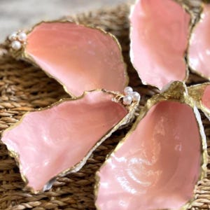 May include: Pink oyster shell dishes with gold trim and pearl accents. The shells are arranged on a woven, natural-toned surface. The shells have a glossy finish and are a light pink color.