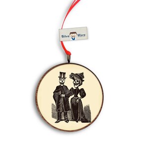 May include: Round wooden ornament featuring a black and white illustration of a skeleton couple in vintage attire. The ornament has a cream-colored background and a red ribbon for hanging. The tag reads "Silva Wars".