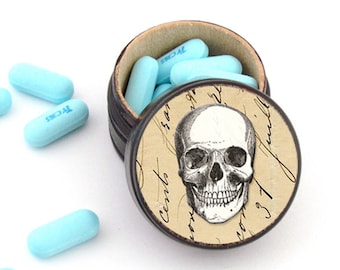 May include: A small, round, black and white pill box with a skull and crossbones design. The box is filled with light blue pills.