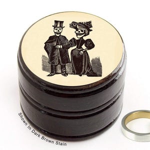 May include: A round wooden ring box with a black stain finish. The lid features a black and white illustration of two skeletons dressed in Victorian attire. The text "Shown in Dark Brown Stain" is printed below the box.