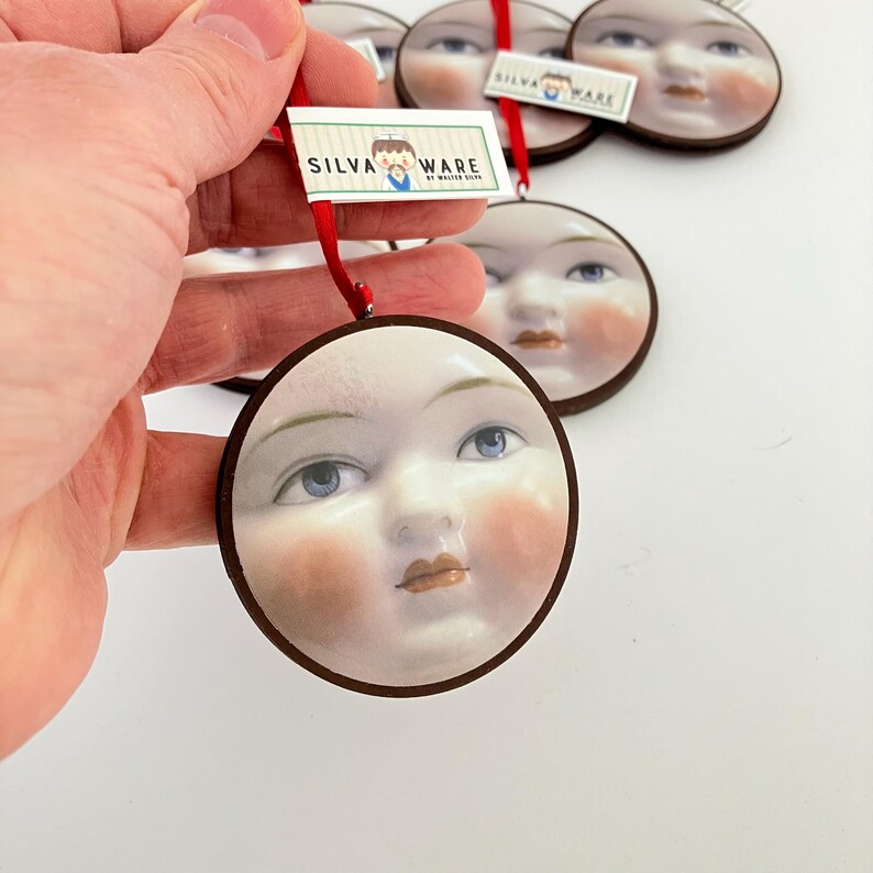 May include: Round ornament featuring a painted face with blue eyes, rosy cheeks, and a brown border. The ornament is held by a red ribbon with a tag that reads "SILVA WARE BY WALTER SOREN". Other similar ornaments are in the background.
