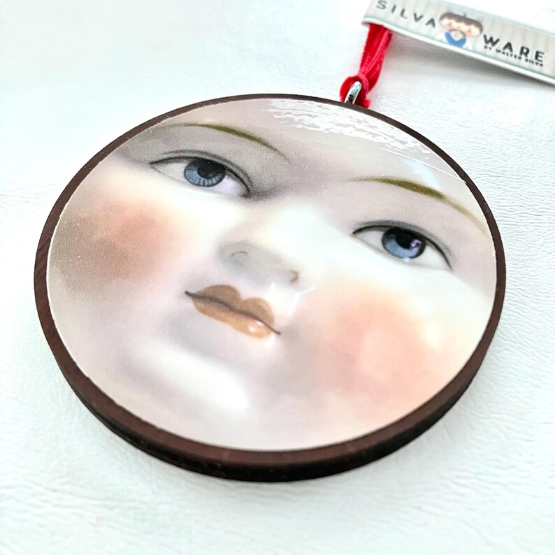 May include: Round ornament featuring a painted face with blue eyes, rosy cheeks, and a brown wooden border. A red ribbon is attached for hanging. The tag reads "SILVA WARE by Walter Shaw."