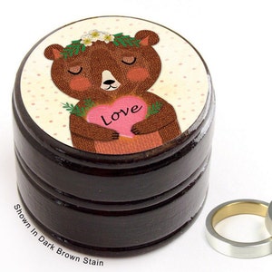 May include: A dark brown stained wooden ring box with a cartoon bear illustration on top. The bear holds a pink heart with the word "Love". Two silver rings with gold interiors are next to the box. Text on the box reads "Shown In Dark Brown Stain".