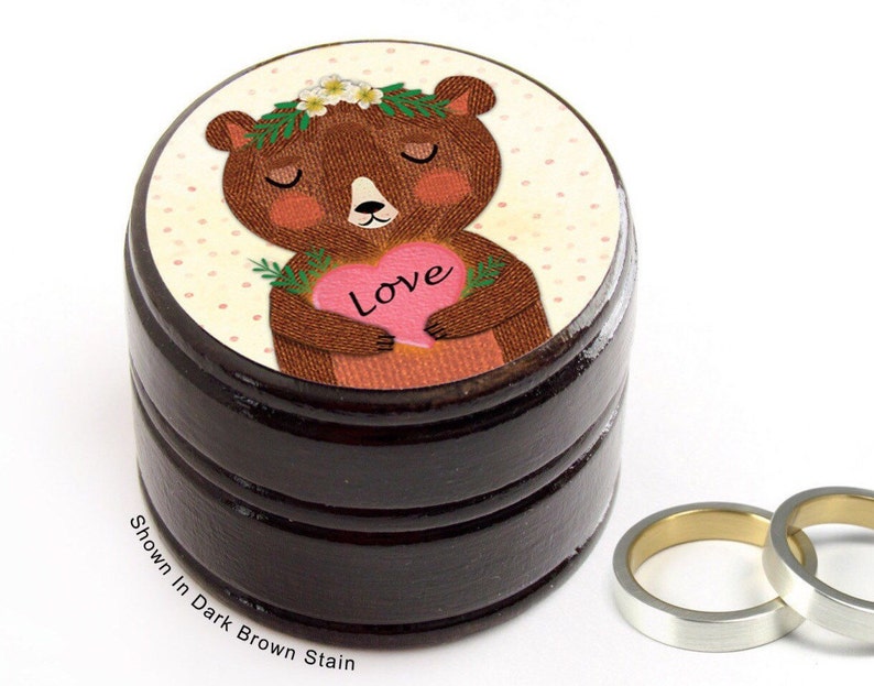 May include: A dark brown stained wooden ring box with a cartoon bear illustration on top. The bear holds a pink heart with the word "Love". Two silver rings with gold interiors are next to the box. Text on the box reads "Shown In Dark Brown Stain".