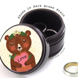 May include: A dark brown wooden ring box with a soft-lined interior. The lid features a cartoon bear holding a pink heart with the word "Love". The box is shown in a dark brown stain.