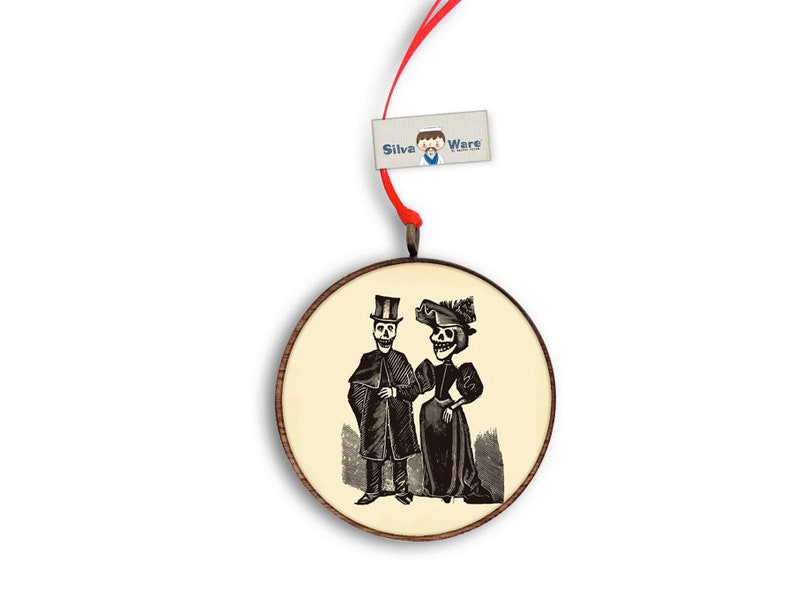 May include: Round wooden ornament featuring a black and white illustration of a skeleton couple in vintage attire. The ornament has a cream-colored background and a red ribbon for hanging. The tag reads "Silva Wars".